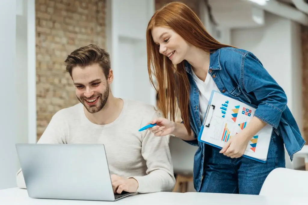 Homme et femme discutant d'un projet professionnel devant un ordinateur portable, avec des graphiques sur un tableau